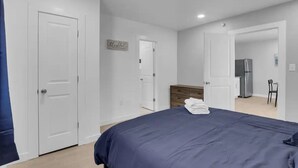 Un dormitorio con cama, cómoda, refrigerador y una silla en una habitación moderna y minimalista con paredes y puertas blancas.