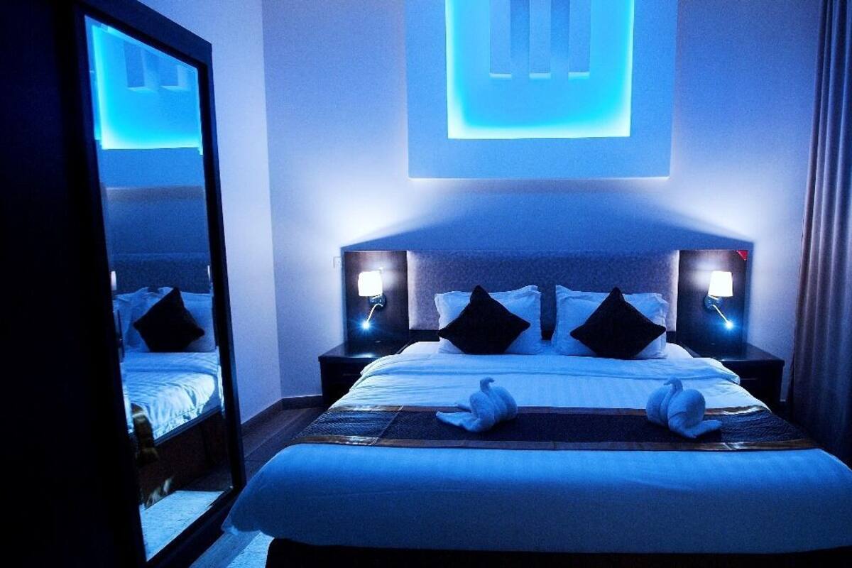 Ein Hotelzimmer mit einem großen Bett, zwei Nachttischen mit Lampen, einem Spiegel und einem wandmontierten Projektor.