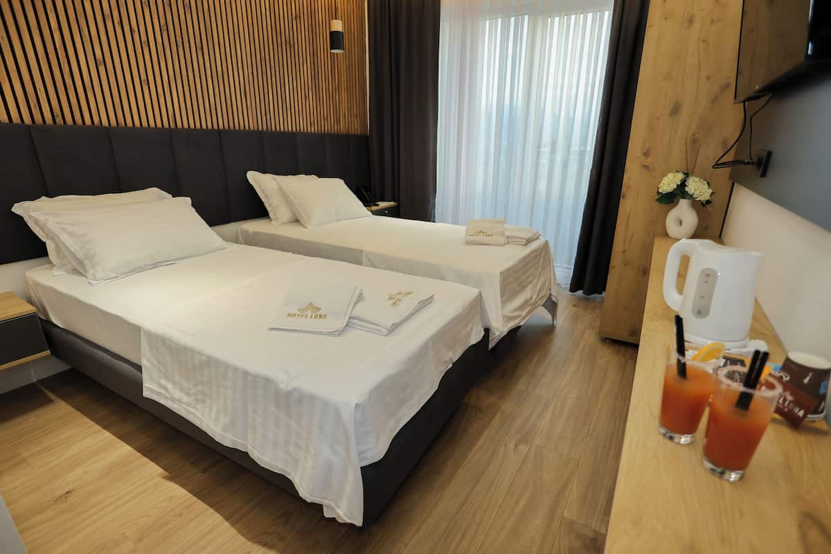 A hotel room with two beds, a wooden headboard, a bedside table with a lamp, a coffee maker, and two drinks on the table.