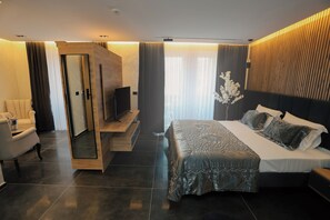 A modern hotel room with a large bed, a seating area with a chair, a TV, and a wooden cabinet. - Hotel Luna (Prizren)