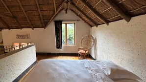 A room with a bed, a wicker chair, a window with curtains, and a wooden ceiling.