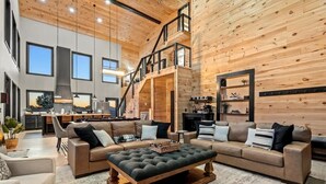 A spacious living area with a large sofa set, a wooden coffee table, and a staircase leading to an upper level. - Luxury Smoky Mtn Cabin | Infinity Pool & Views (Cosby)