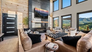 Un salon moderne avec des canapés en cuir, une cheminée, un téléviseur et de grandes fenêtres offrant une vue sur les montagnes.