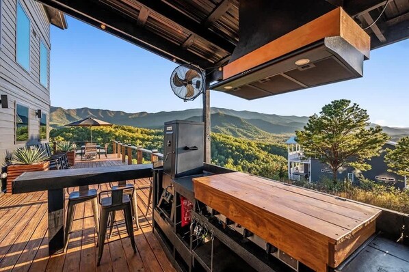 An outdoor deck with a grill, bar stools, and a wooden table overlooking a mountainous landscape. - Luxury Smoky Mtn Cabin | Infinity Pool & Views (Cosby)