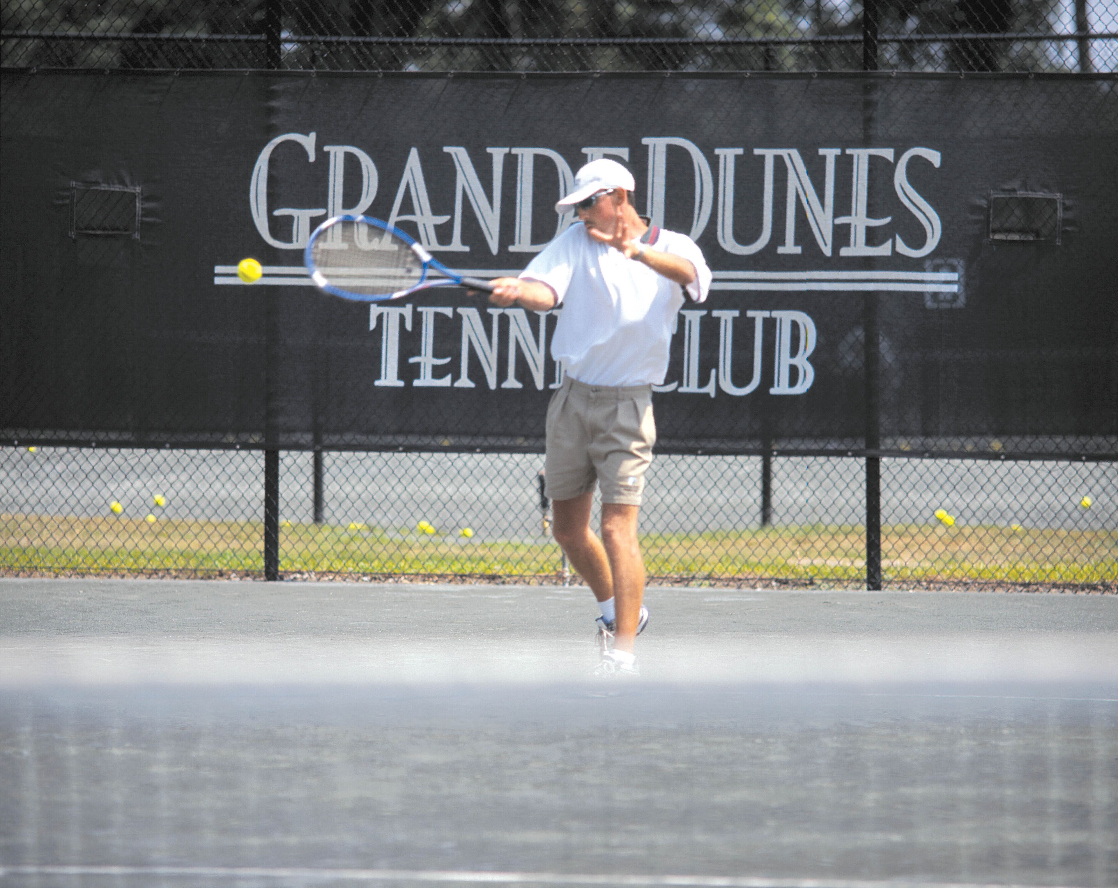 Ein Tennisplatz mit Zaun und einem Banner mit der Aufschrift 'GRAND DUNES TENNIS CLUB'.