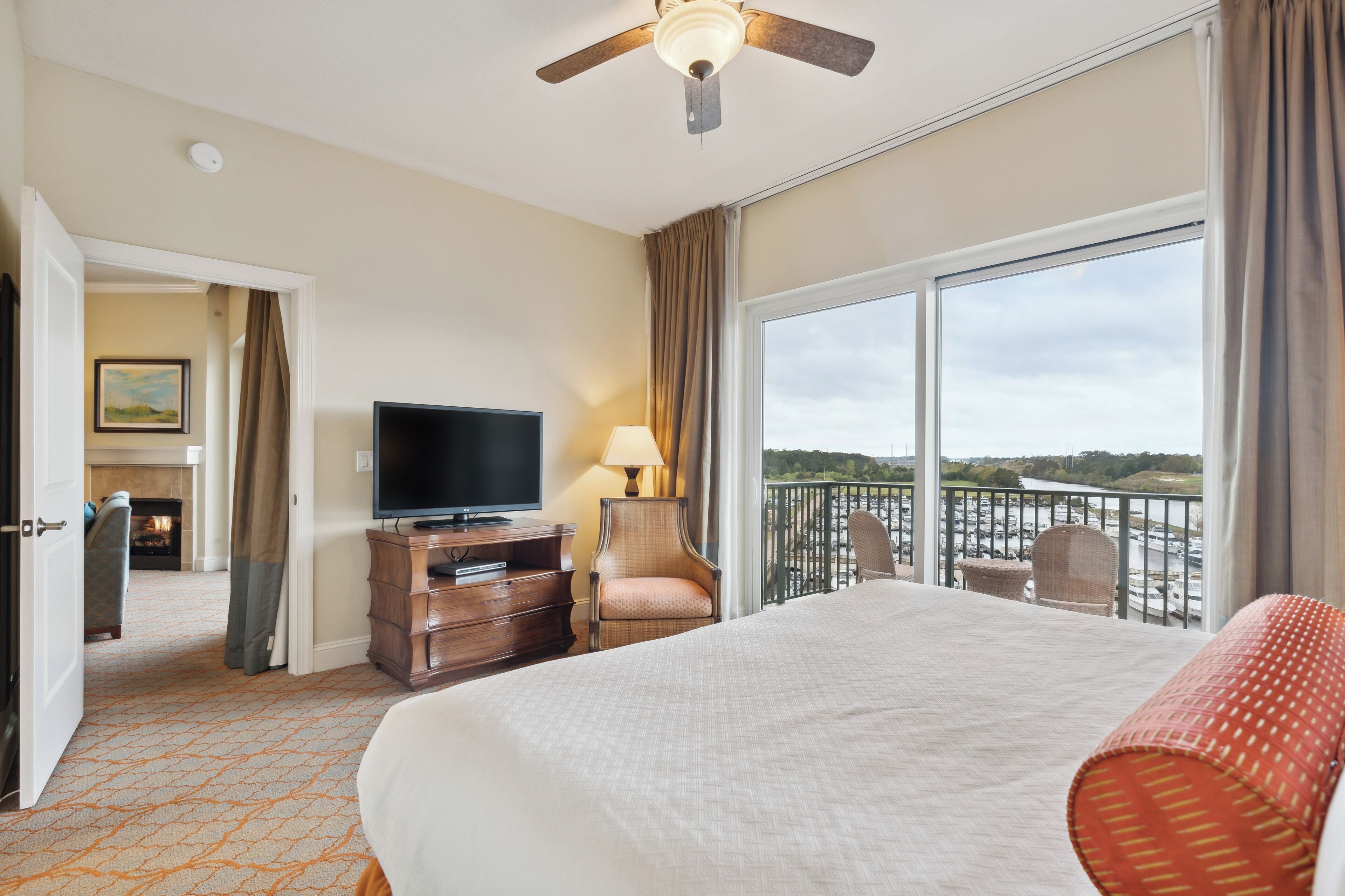 A hotel room with a large bed, a television, a desk with a lamp, a chair, and a balcony with a view of the outdoors.