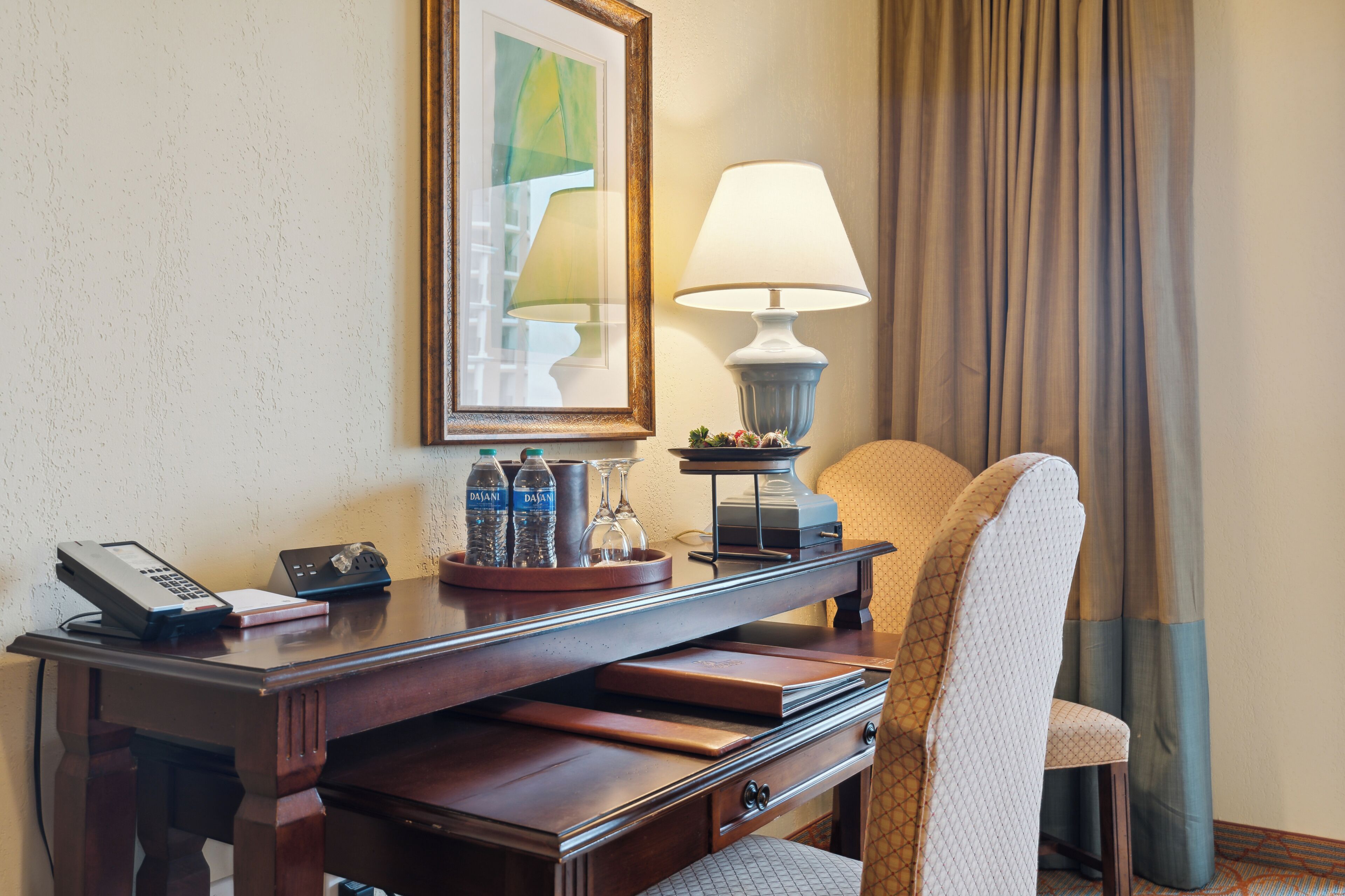Hotelkamer met een houten bureau, een lamp, een stoel, een ingelijste foto en een raam met gordijnen.