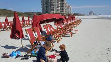 A beach with rows of red beach chairs and umbrellas, a tall building in the background, and a clear sky.