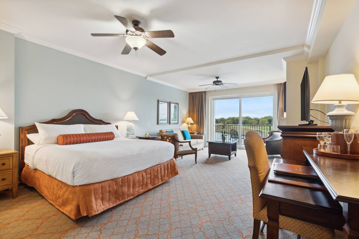 A hotel room with a large bed, a desk, a chair, a balcony with a view, and two ceiling fans.