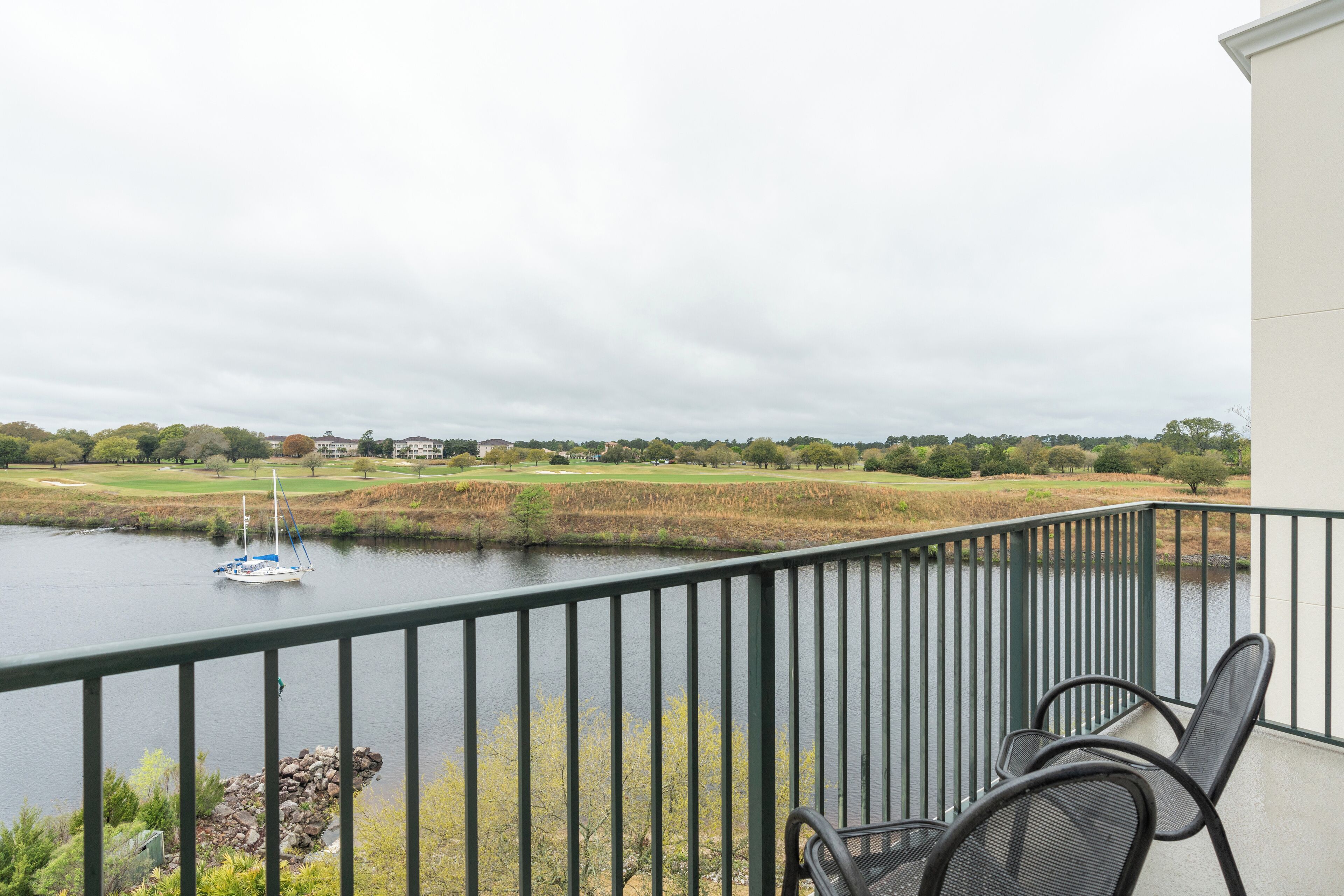 A balcony with a view of a lake, a sailboat, and a golf course in the background.