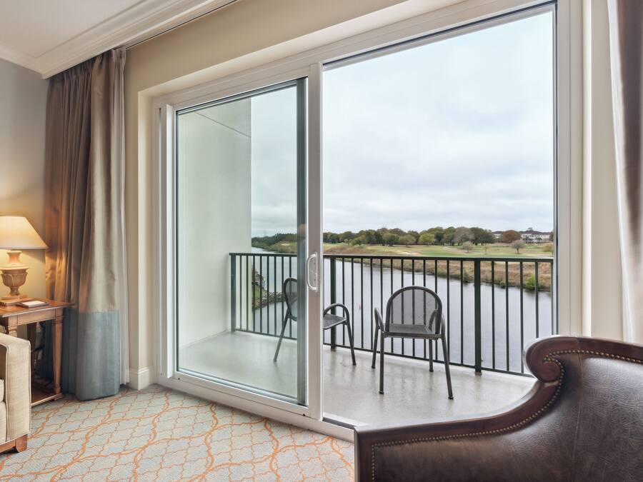 A hotel room with a balcony, a sofa, a table with a lamp, and a view of a golf course.