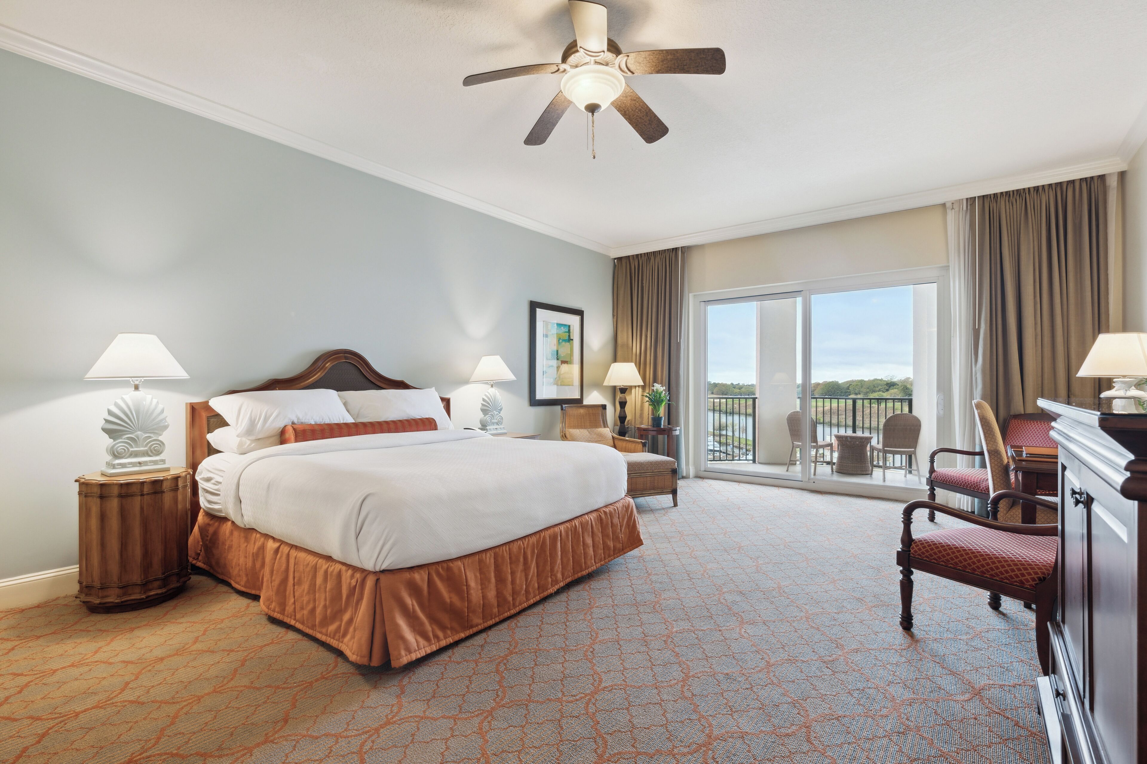 A hotel room with a large bed, two bedside tables with lamps, a ceiling fan, a balcony with chairs, and a view of the outdoors.