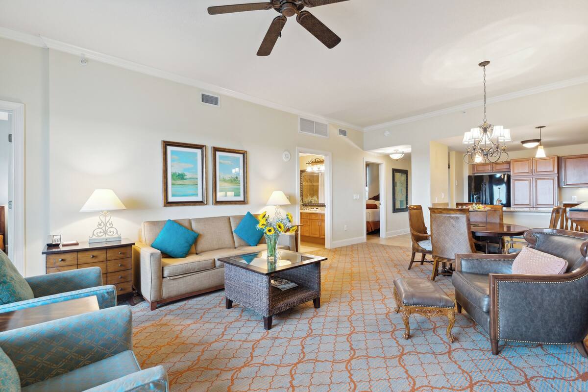 A spacious living room with a sofa, armchairs, a coffee table, and a chandelier. There is a dining area with a table and chairs, and a kitchen in the background.