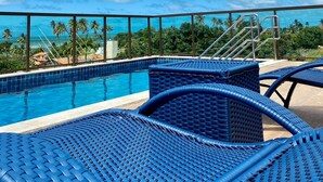 A poolside area with blue lounge chairs, a clear blue sky, and a view of palm trees and buildings in the background. - Green Porto 104  (Ipojuca)