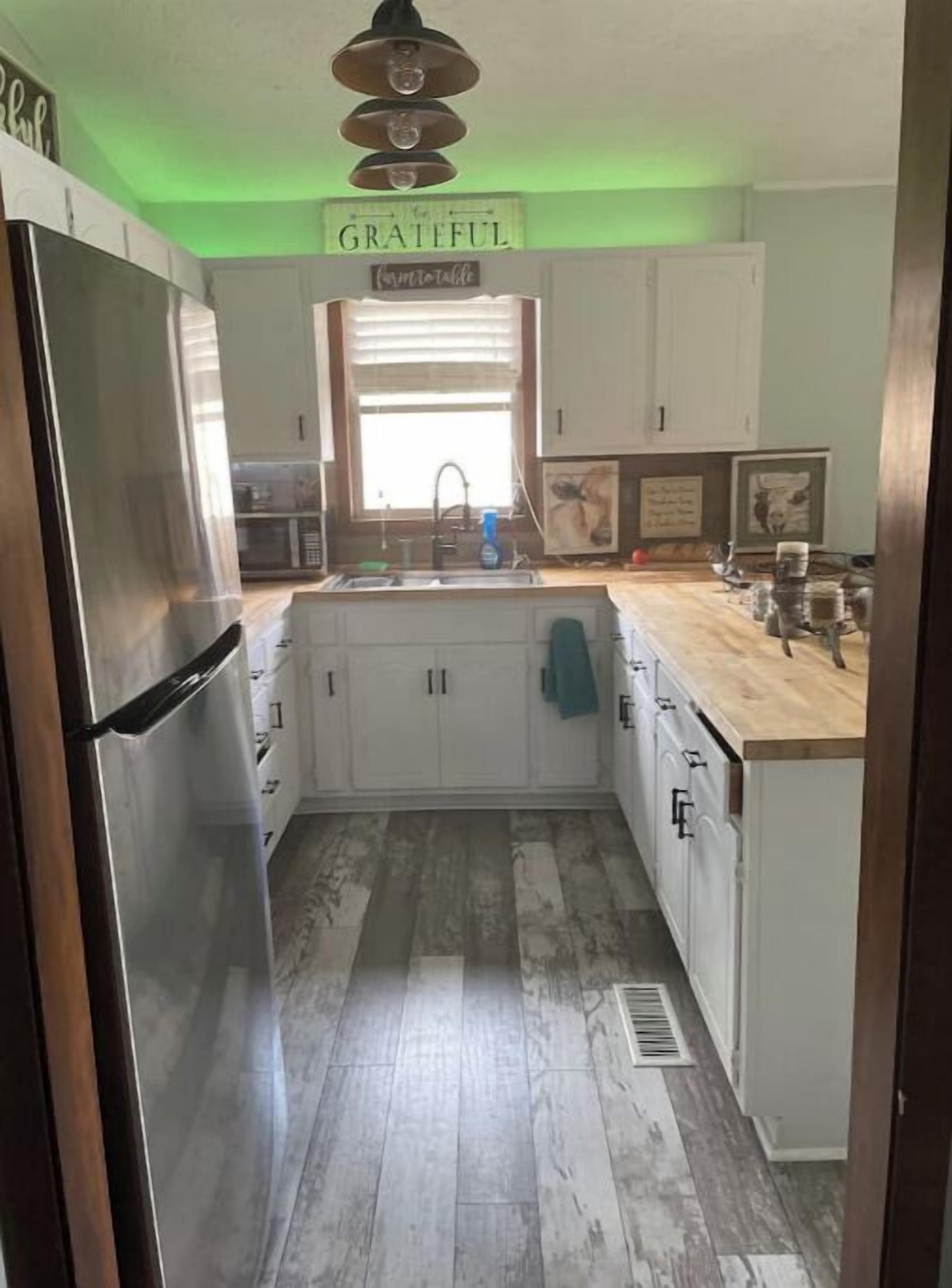 A kitchen with white cabinets, a stainless steel refrigerator, a window with blinds, and a sign that says "GRATEFUL".