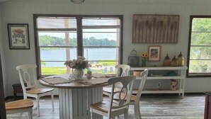 Un espace repas avec une table ronde et des chaises blanches, de grandes fenêtres offrant une vue sur la verdure et l’eau, et un plafond en bois.