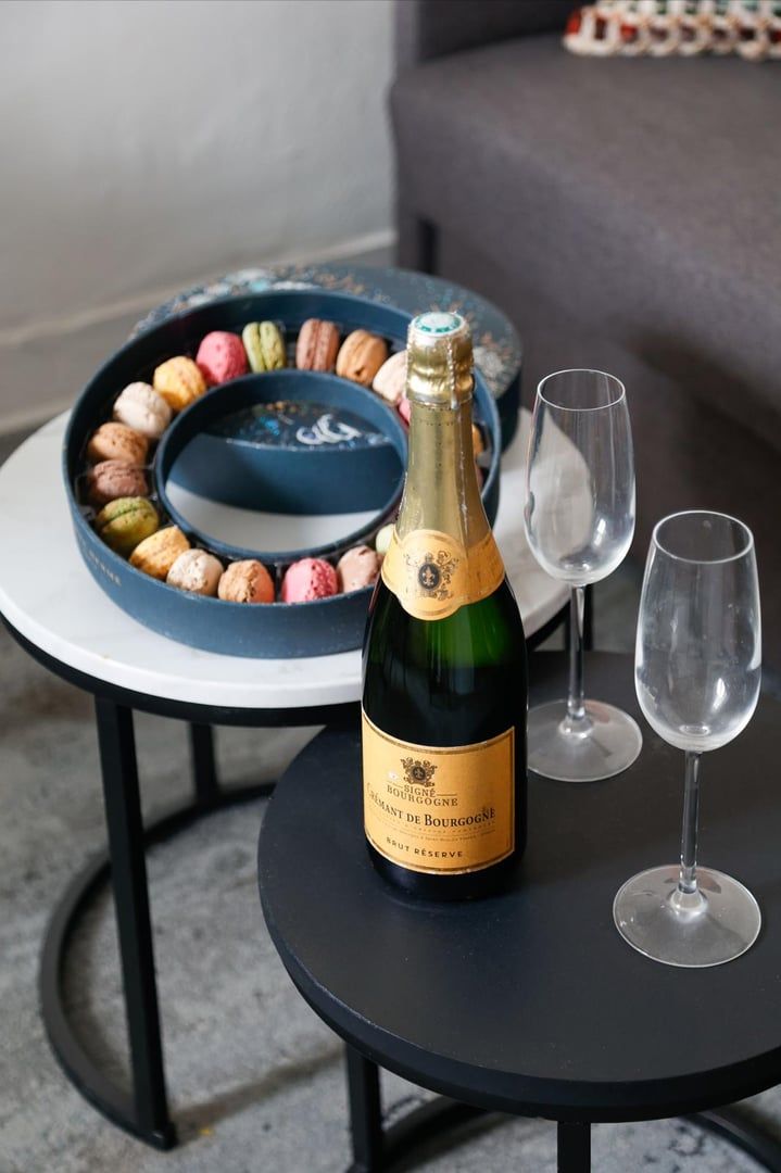 Une bouteille de champagne, deux verres et une assiette remplie de macarons multicolores sur une table ronde.