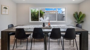 A dining area with a rectangular table, four black chairs, a vase with flowers, and a view of a residential area through a window. - Girls  & Family Trip+Pool+beauty bar, San Diego (La Mesa)