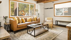 A cozy living room with a leather sofa, a wooden coffee table, and large windows offering a view of a cabin. - National Forest View Chalet: Hot Tub, A/C & BBQ (Big Bear Lake)