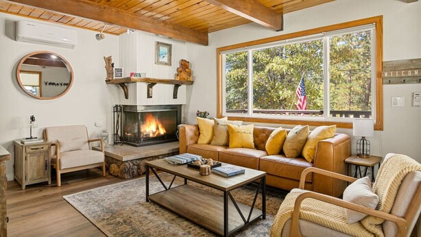 A cozy living room with a fireplace, wooden ceiling beams, a leather sofa, and a large window with an American flag. - National Forest View Chalet: Hot Tub, A/C & BBQ (Big Bear Lake)