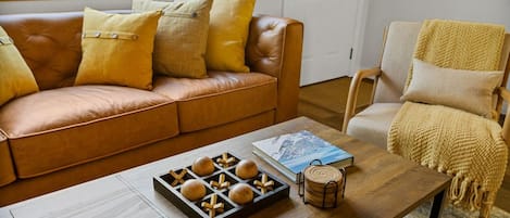Una sala de estar con un sofá de cuero marrón, una mesa de centro de madera con un juego de mesa, un libro y una cesta tejida.