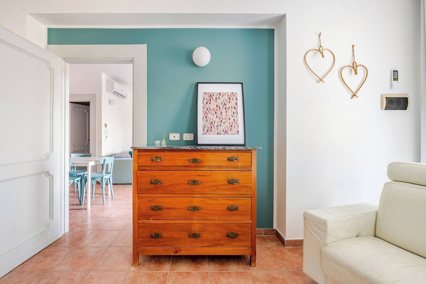 Una stanza con una parete verde acqua, un comò in legno con maniglie in ottone, un quadro incorniciato e un divano bianco.
