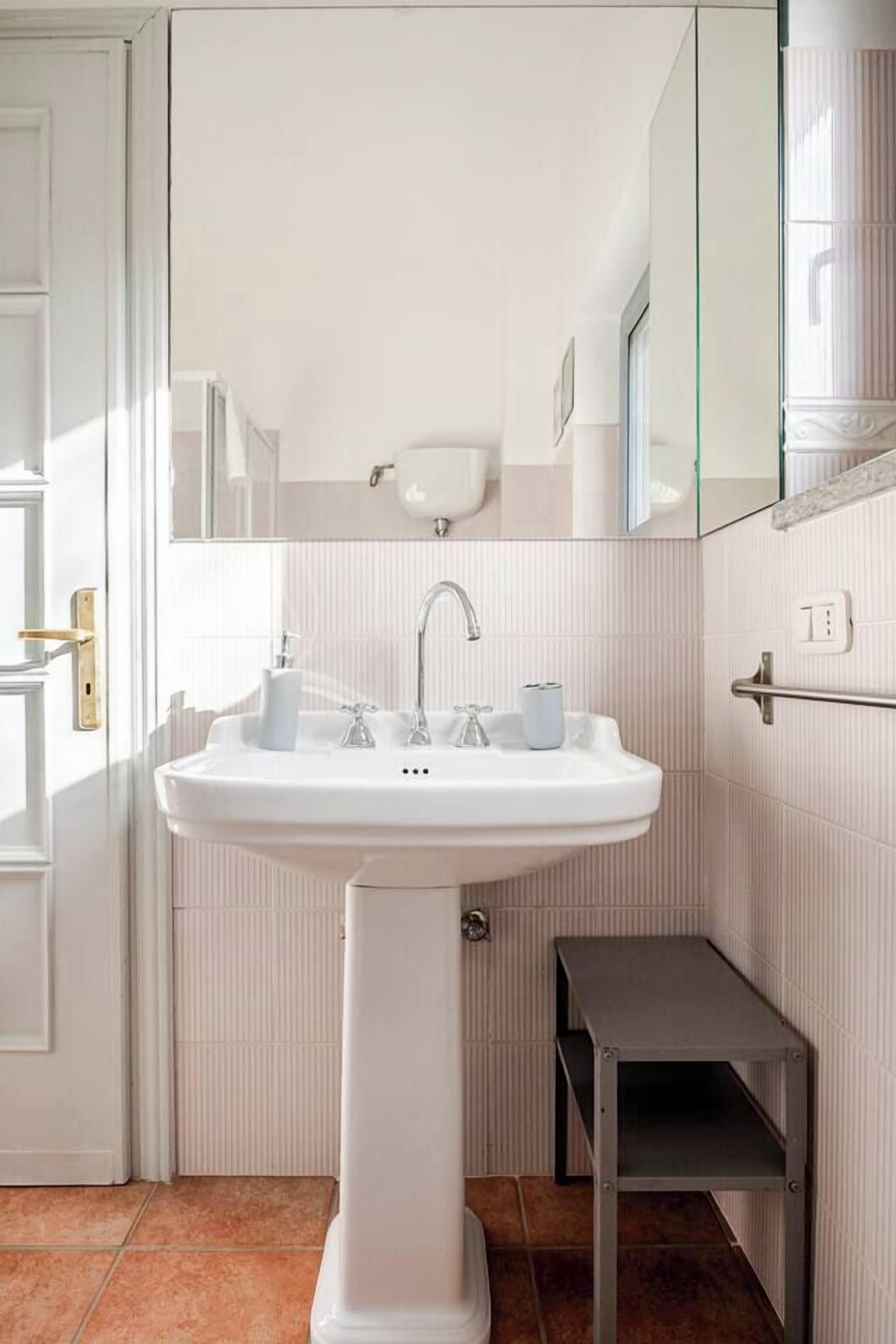 A bathroom with a pedestal sink, a mirror, and a small shelf.