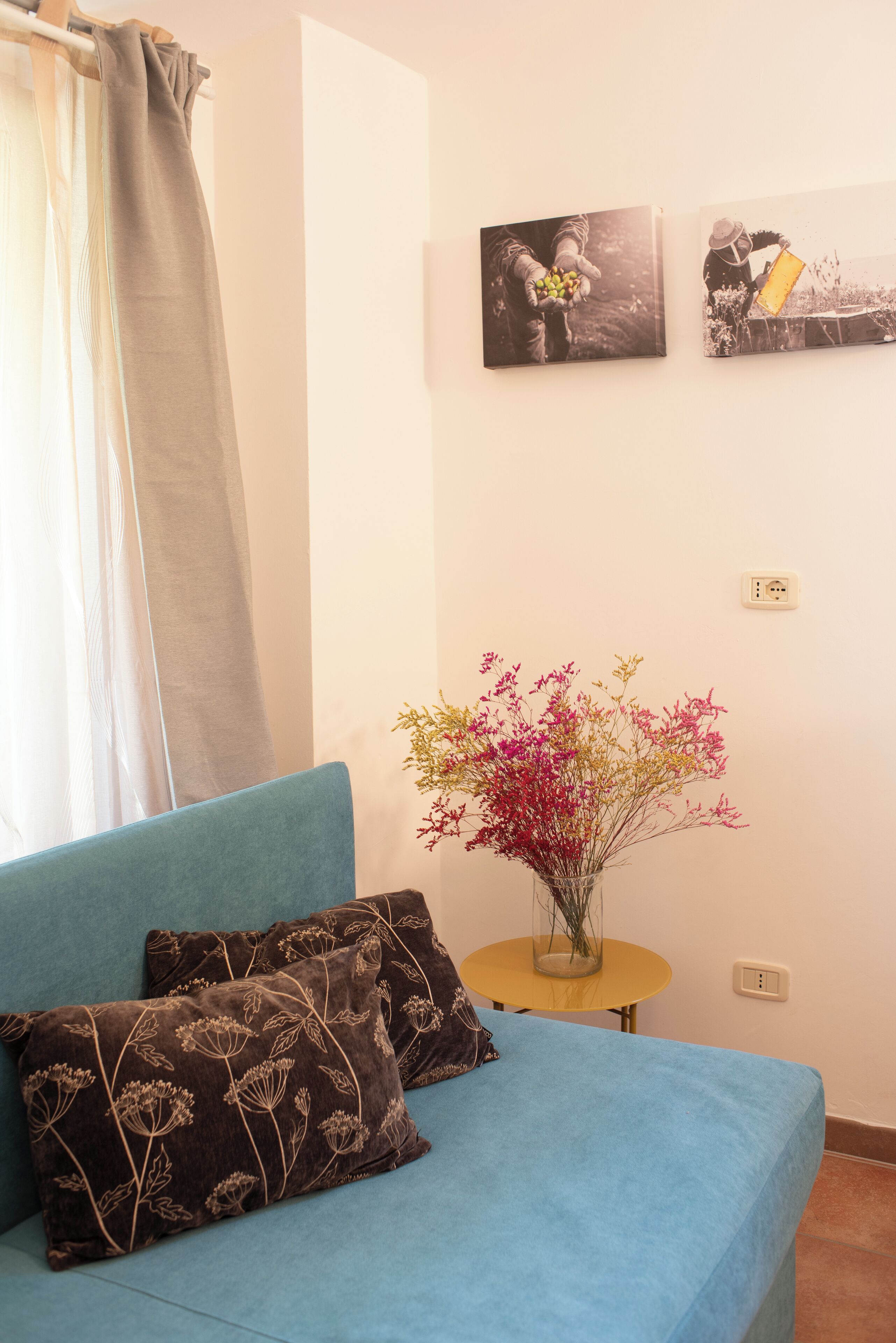 Une pièce avec un canapé bleu, deux coussins à motifs, une petite table ronde ornée d’un vase de fleurs et deux photographies en noir et blanc accrochées au mur.