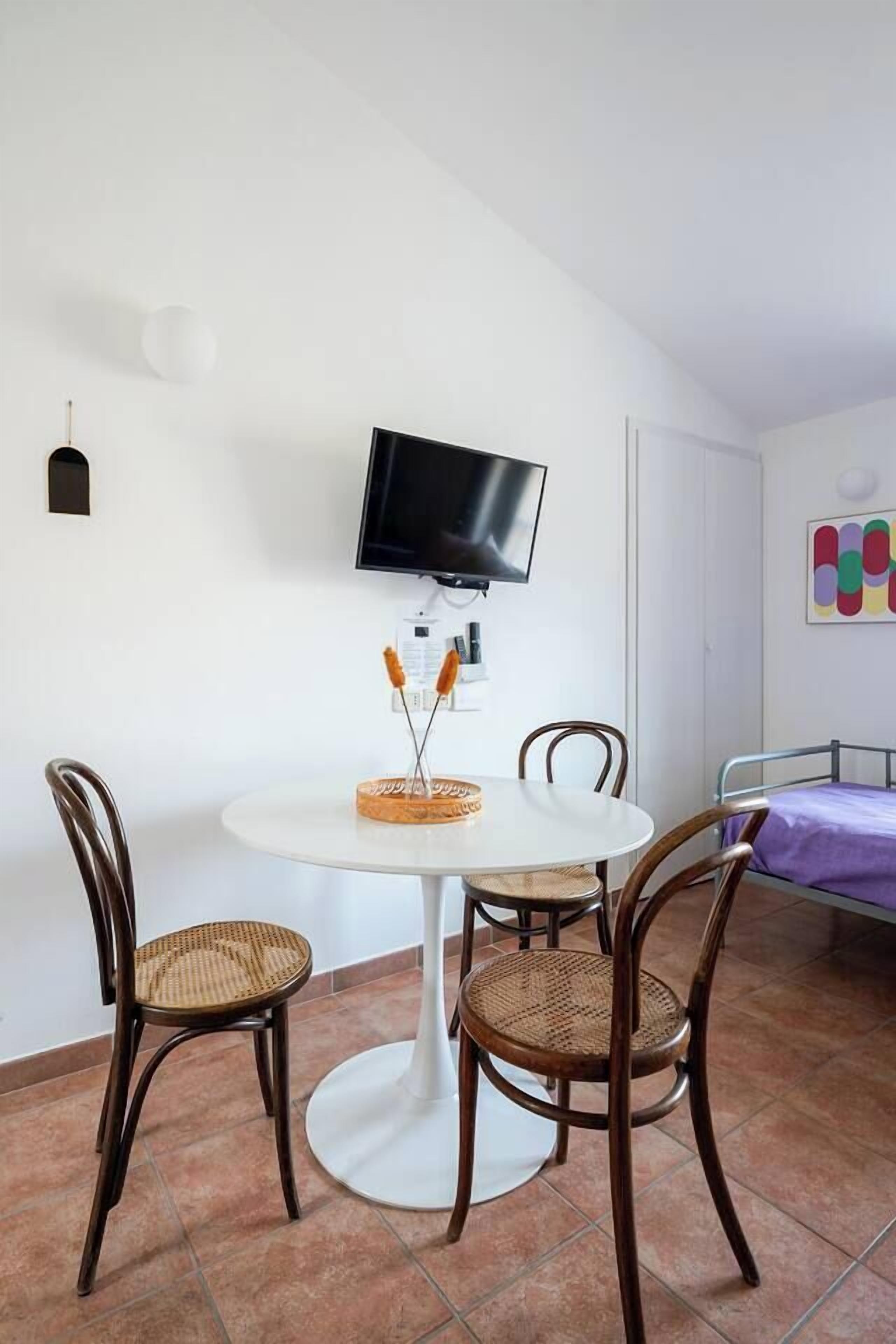 Une petite pièce bien éclairée, avec une table de salle à manger ronde, deux chaises en osier, un téléviseur fixé au mur et un lit recouvert d’une couverture violette.