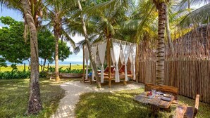 Ein tropischer Außenbereich mit Sitzgelegenheit, einem Holztisch und Stühlen, umgeben von Palmen und Blick aufs Meer.