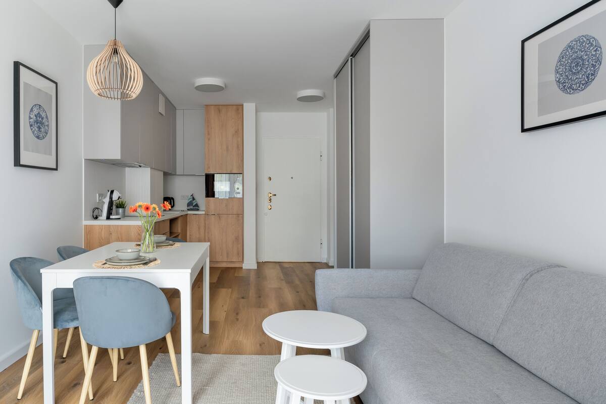 A modern living room with a grey sofa, a white dining table with blue chairs, and a kitchen in the background.