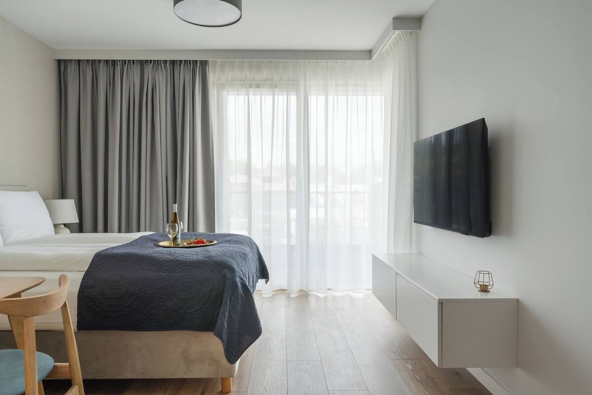 A hotel room with a bed, a bedside table, a TV mounted on the wall, and a window with curtains.