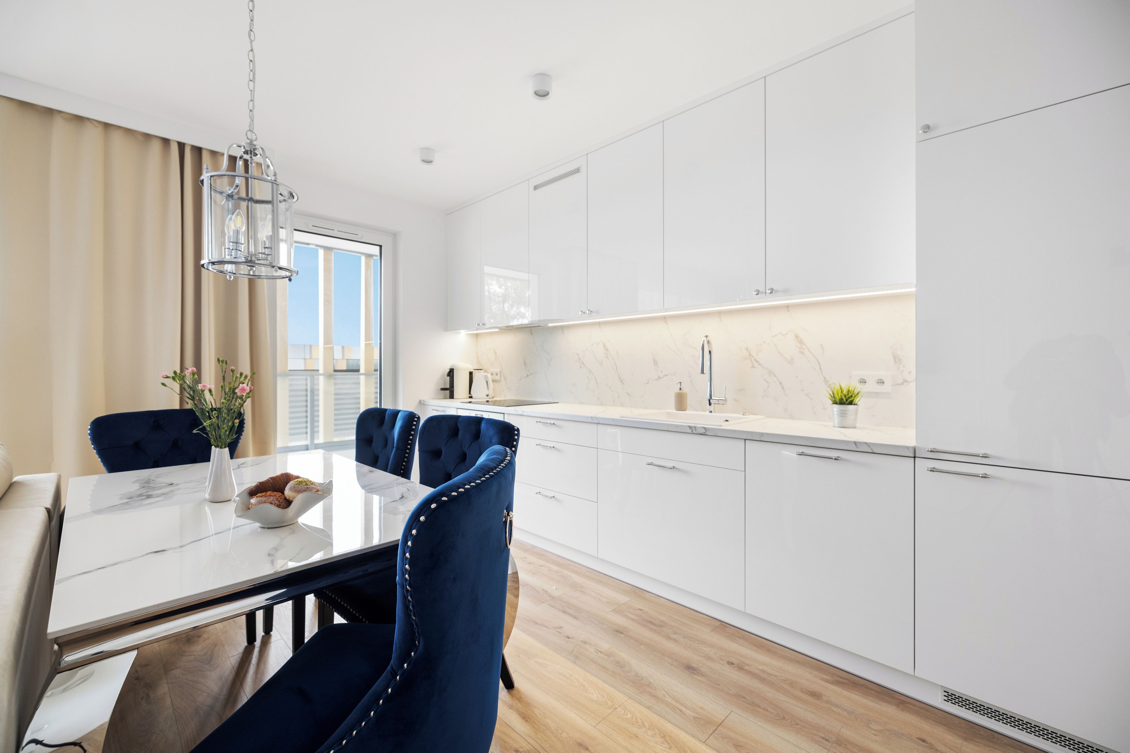 A modern kitchen with white cabinets, a marble countertop, and a dining area with a white table and blue upholstered chairs.