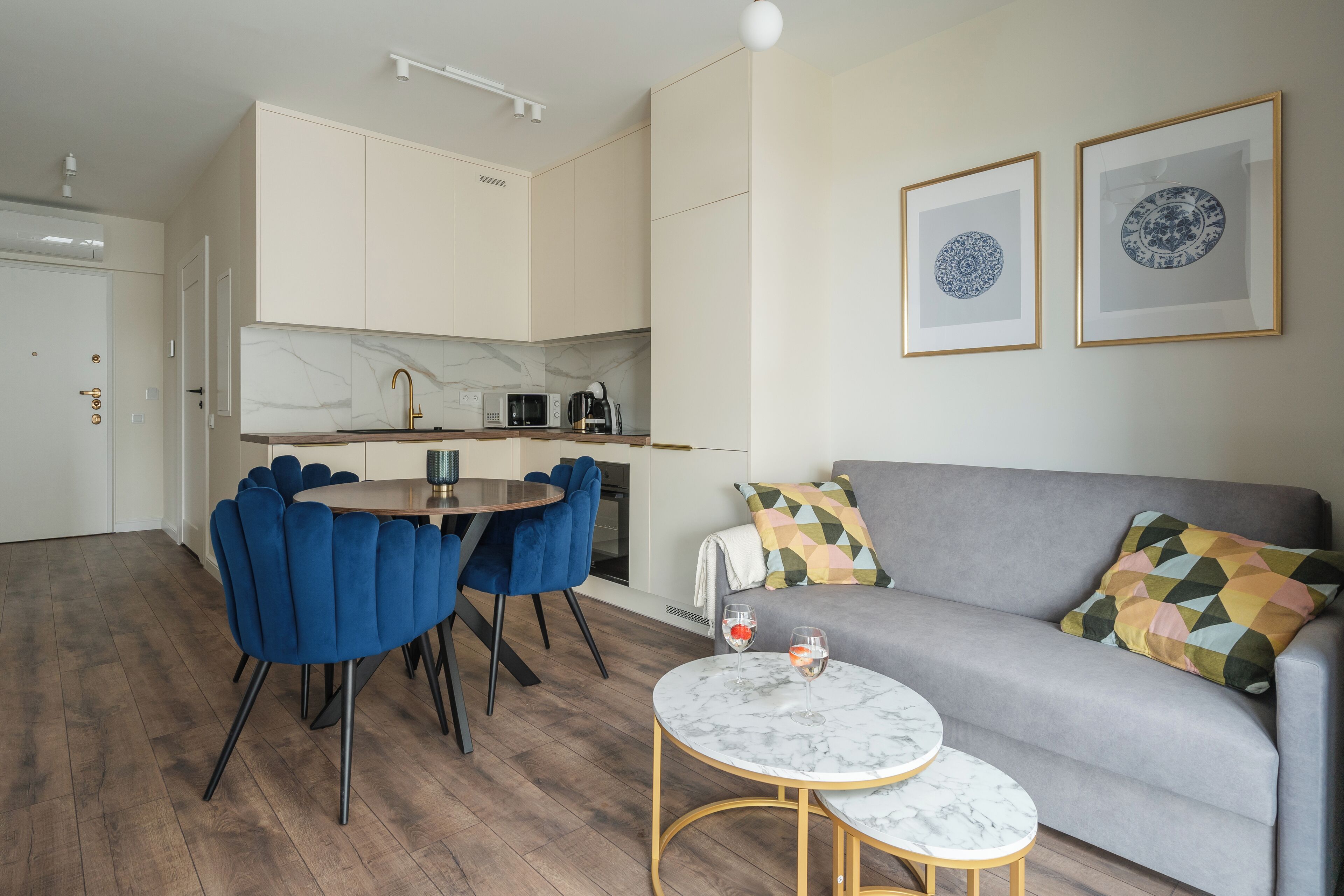 Uma sala de estar moderna com um sofá cinzento, cadeiras azuis, uma mesa de centro de mármore e uma cozinha ao fundo.