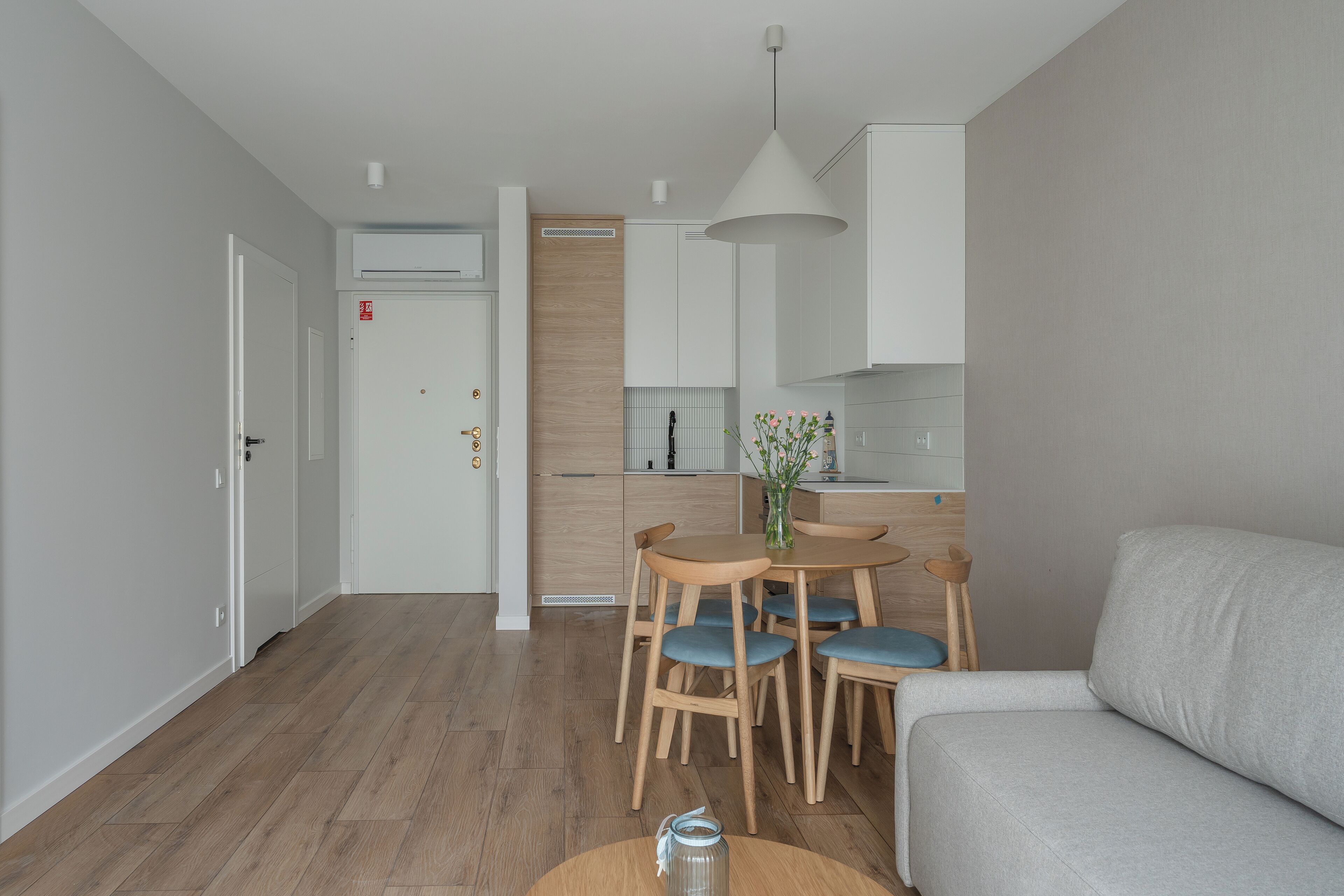 Una sala moderna con un sofá gris, una mesa de comedor de madera con sillas azules y una zona de cocina con armarios blancos y una pared de acento de madera.