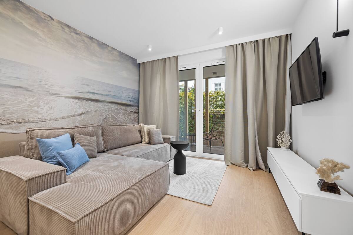 A modern living room with a large sectional sofa, a flat-screen TV mounted on the wall, and a view of the outdoors through large sliding doors.