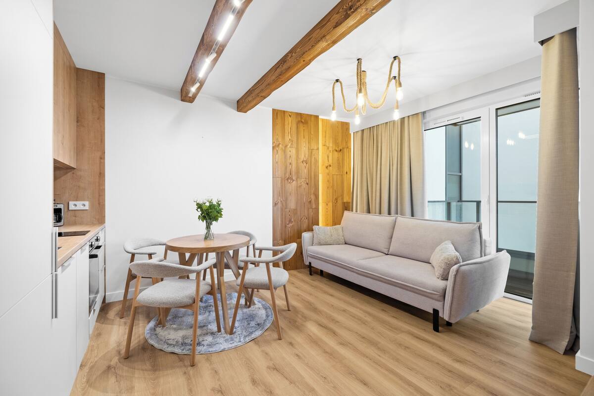 A modern living room with a sofa, dining table, and chairs, wooden flooring, and a large window with curtains.