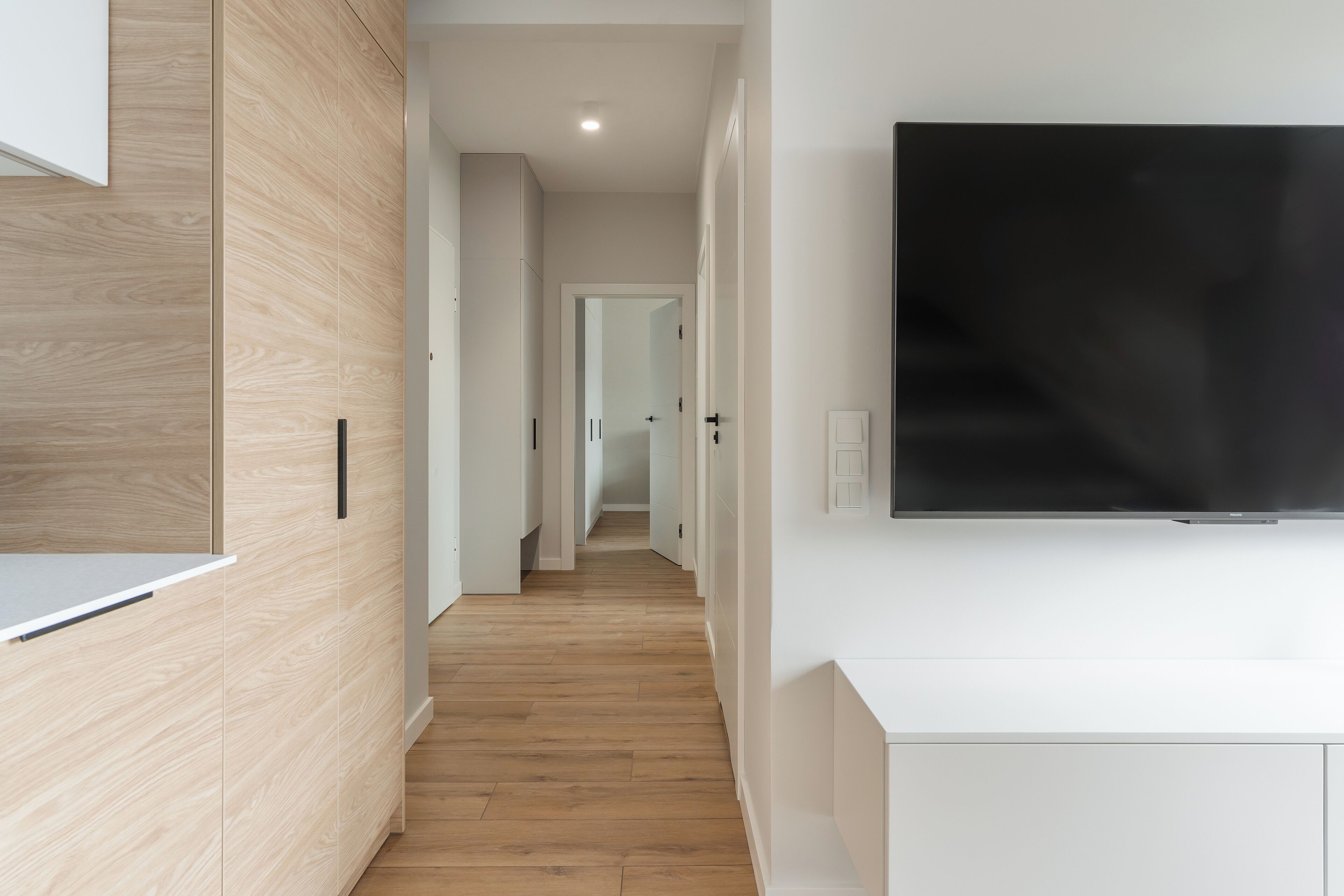 Un pasillo moderno con suelo de madera, paredes blancas y un televisor de pantalla plana montado.