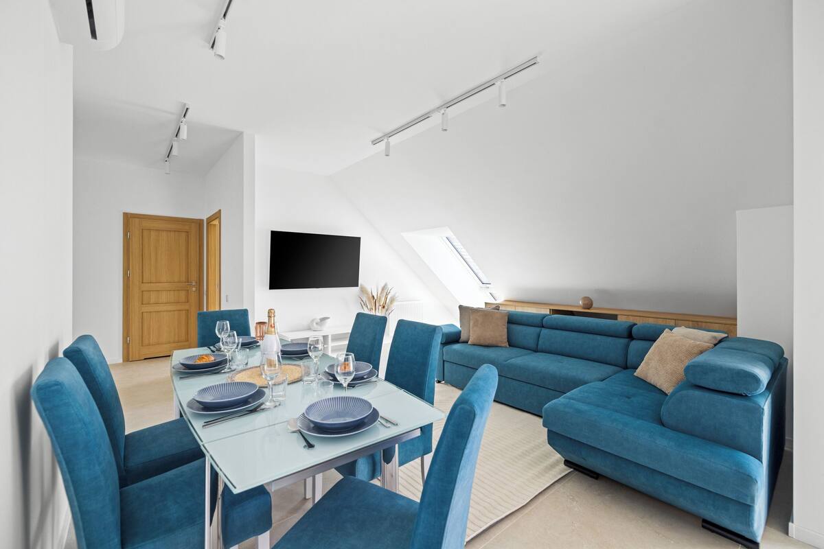 A modern living room with a dining area, featuring a sofa, a dining table set with plates and glasses, and a flat-screen TV mounted on the wall.