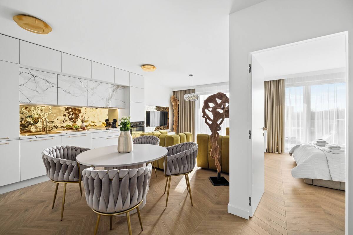 A modern hotel room with a wooden floor, a bed with white linens, a dining area with a round table and upholstered chairs, and a living area with a sofa and a decorative wall piece.