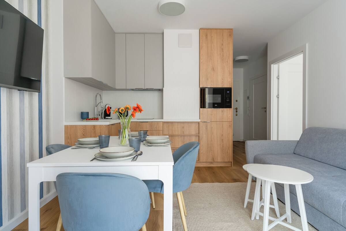 A modern kitchen with white cabinets, a microwave, and a sink. There is a dining table with blue chairs and a vase of flowers. A grey sofa and a small white table are in the living area.