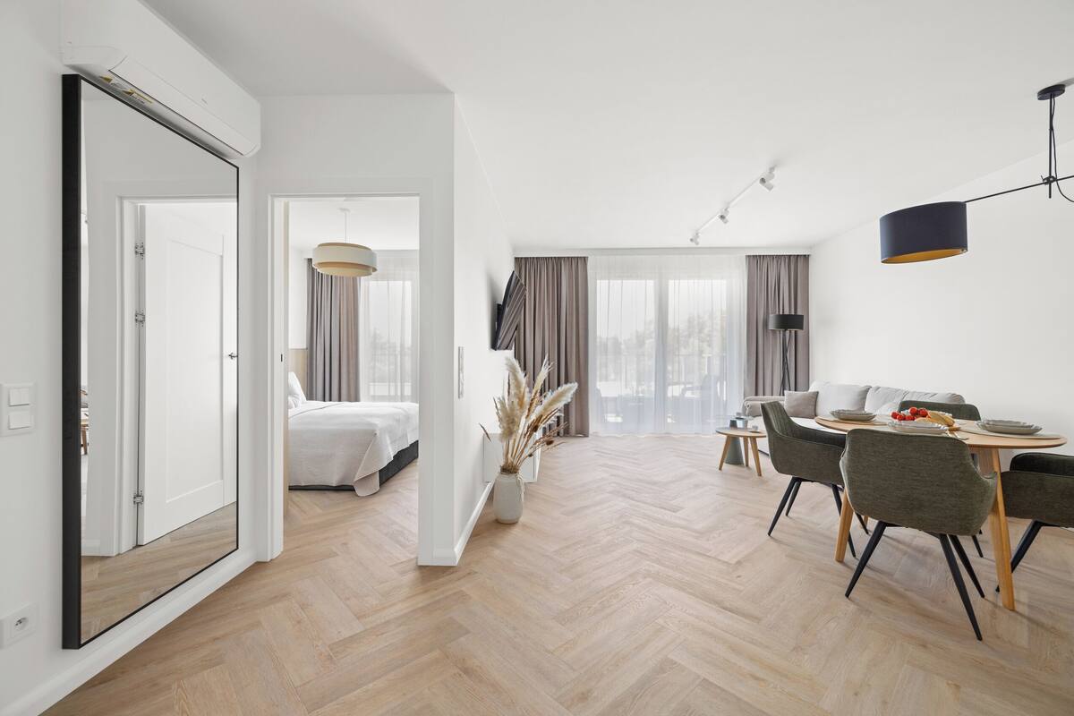 A modern hotel room with a large bed, a dining area with a table and chairs, and a view of the outdoors through large windows.