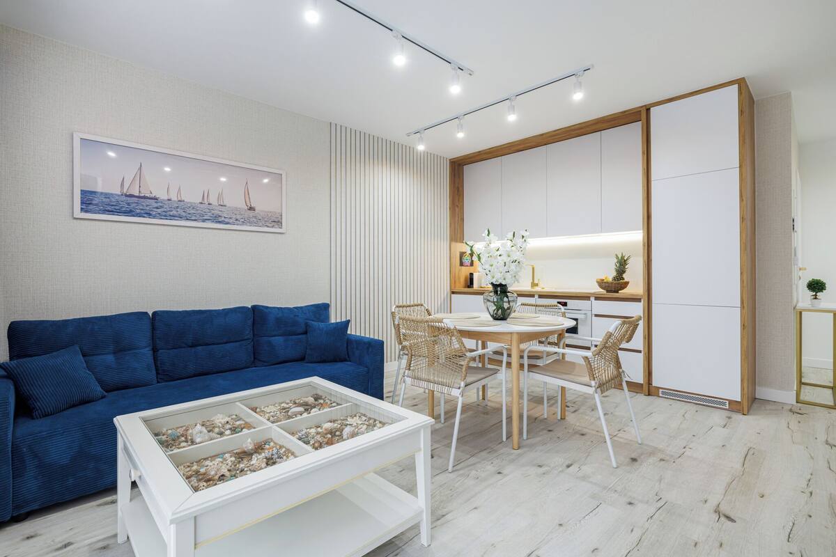 A modern living room with a blue sofa, white coffee table, and a dining area with a wooden table and chairs.