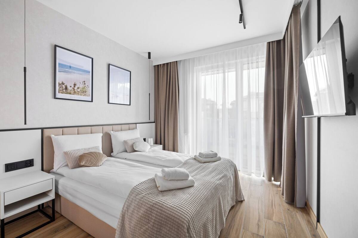 A hotel room with two beds, a TV, a desk, and a large window with curtains.