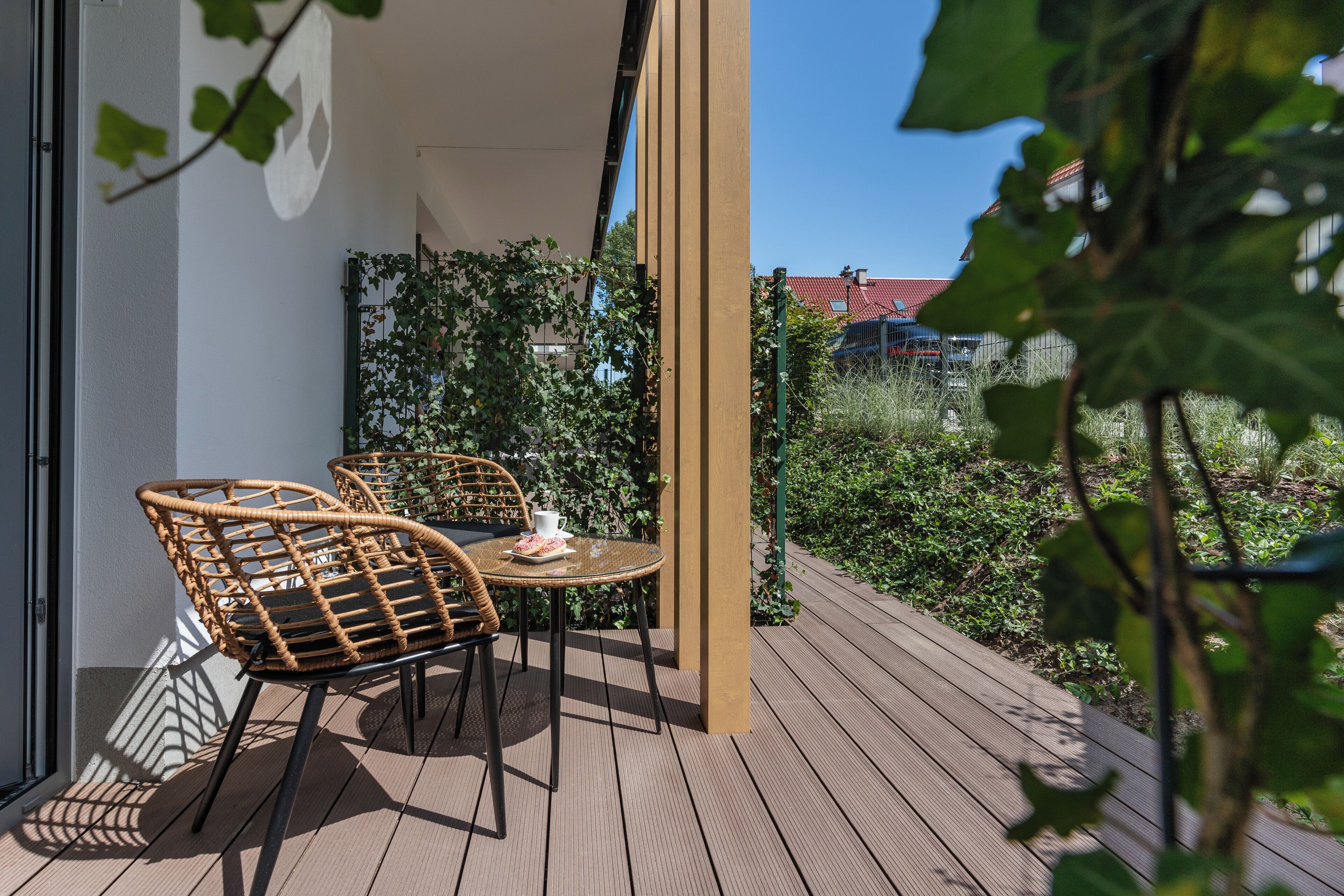 Eine Holzterrasse mit Korbstühlen und einem Tisch, umgeben von viel Grün und blauem Himmel.
