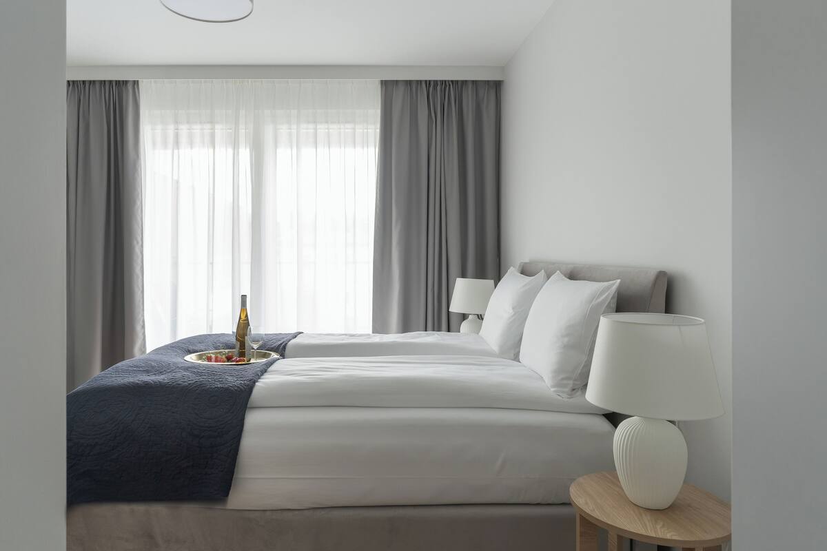 A modern hotel room with a large bed, two bedside lamps, a bedside table, and a view through sheer curtains.