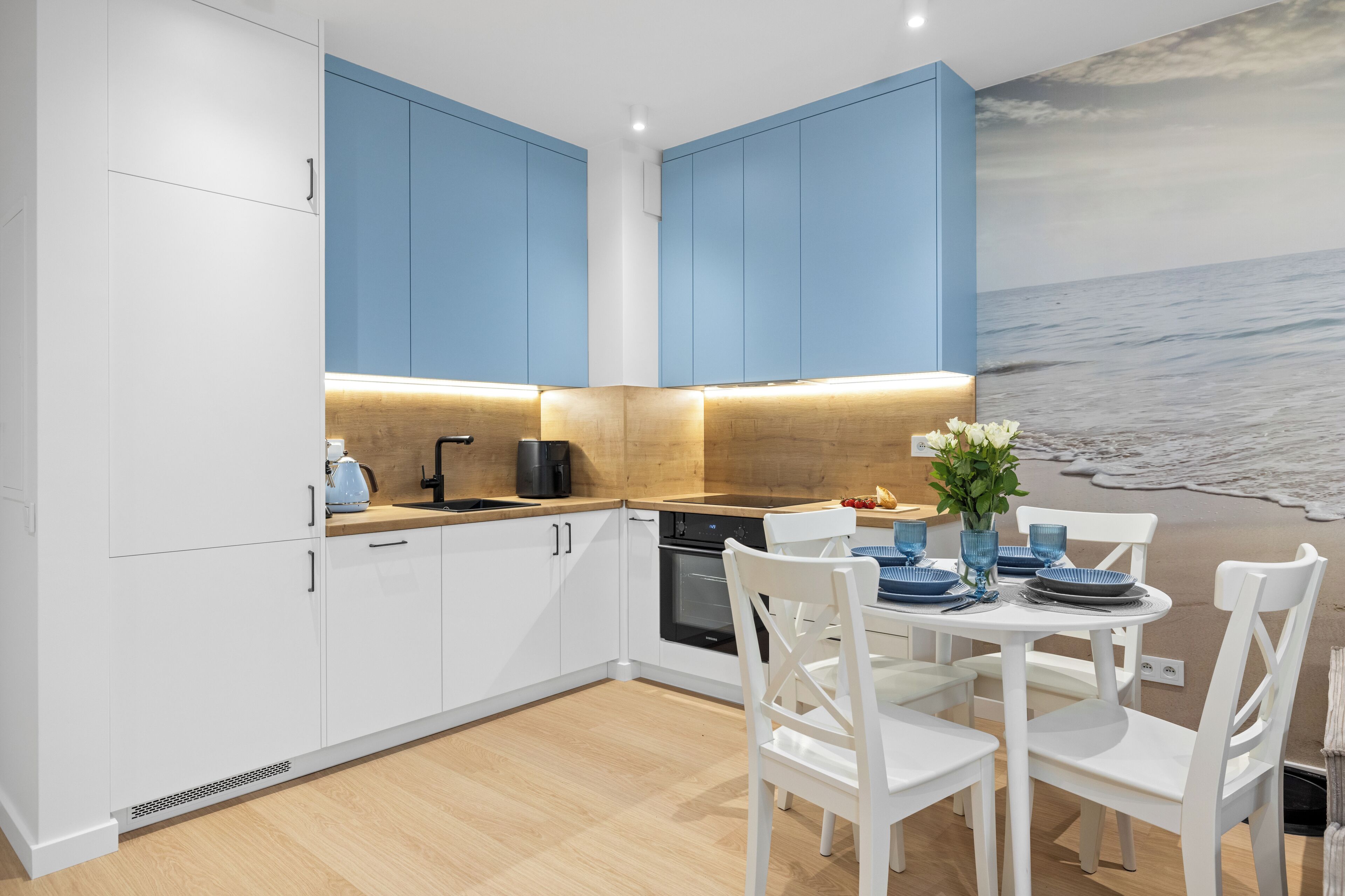 Een moderne keuken met witte en blauwe kastjes, een houten aanrecht en een eethoek met witte stoelen en een tafel.
