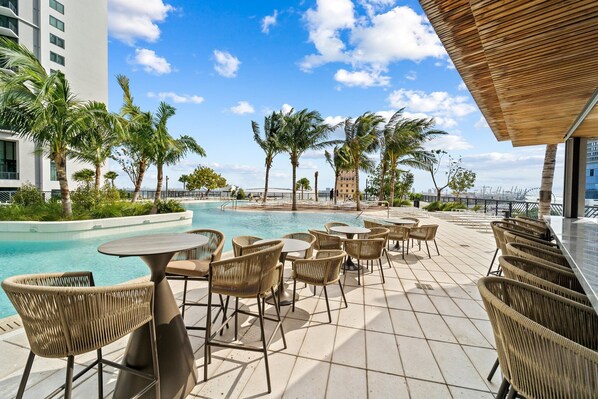 A rooftop pool area with lounge chairs and tables, surrounded by palm trees and modern buildings.