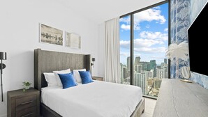 A modern hotel room with a large bed, a nightstand, a desk, and a city view through large windows.