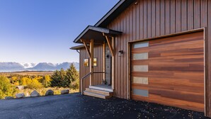 Un bâtiment moderne en bois, doté d’un garage, d’un escalier menant à l’entrée et offrant une vue sur les montagnes en arrière-plan.